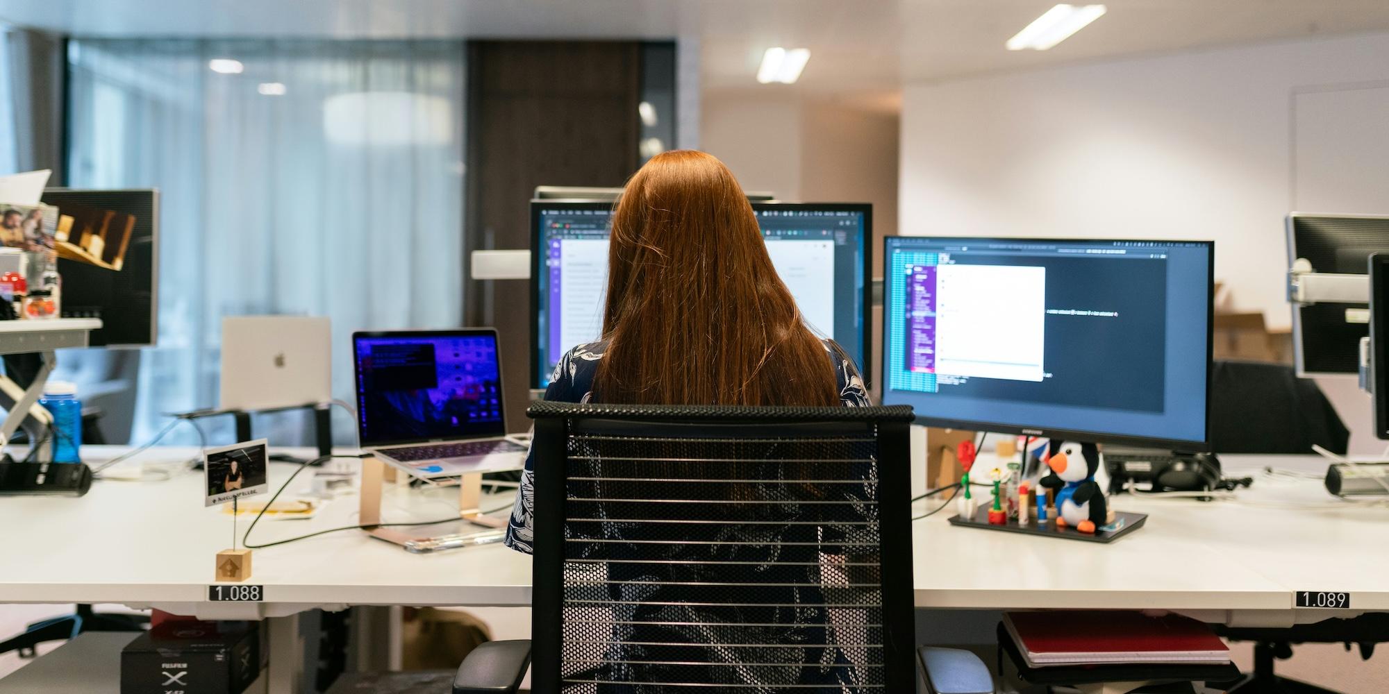 Cover image for this post: A person with long hair is seated at a desk in an office with multiple monitors in front of them with code on the screens. They are viewed from the back.