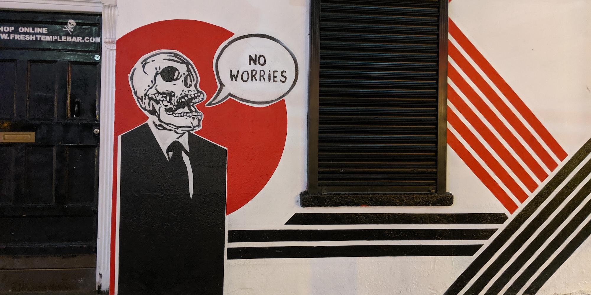 Cover image for this post: Graffiti in London, a skeleton in a suit on, encircled in a red background, says 'No worries'.