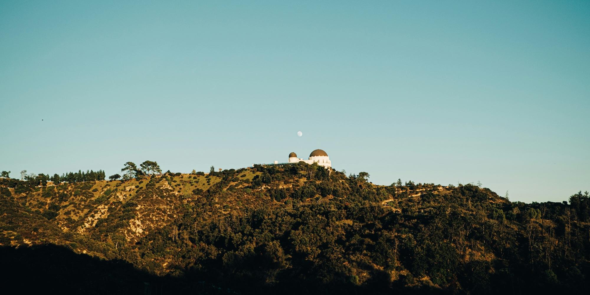 Cover image for this post: Griffith Observatory in LA pictured from afar, with the moon visible in the background during the daytime.