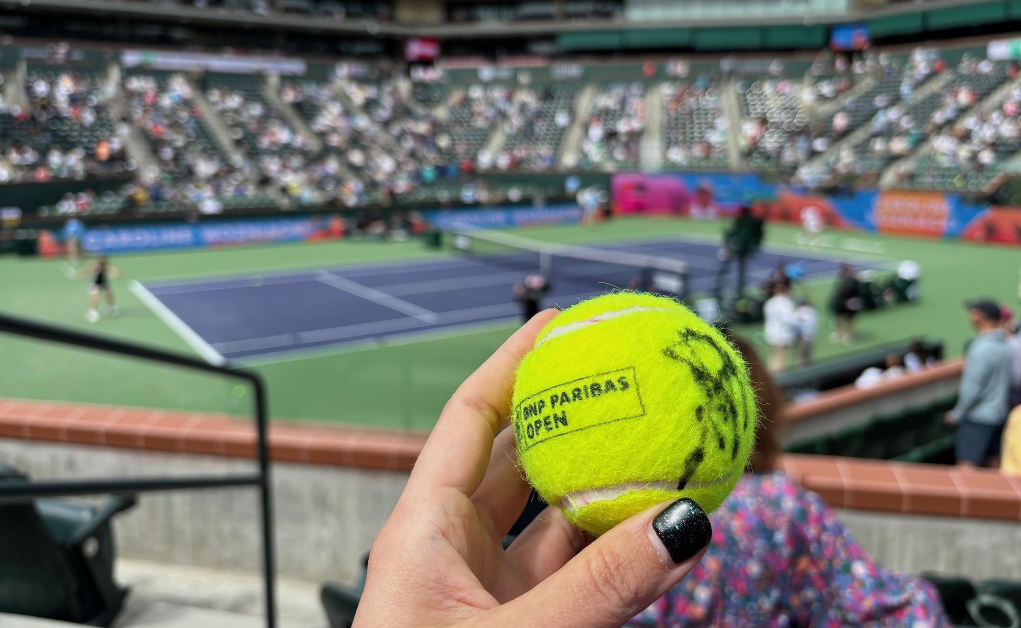 Cover image for this post: A close-up of a tennis ball printed with 'BNP Paribas Open', with center court at Indian Wells blurred in the background.