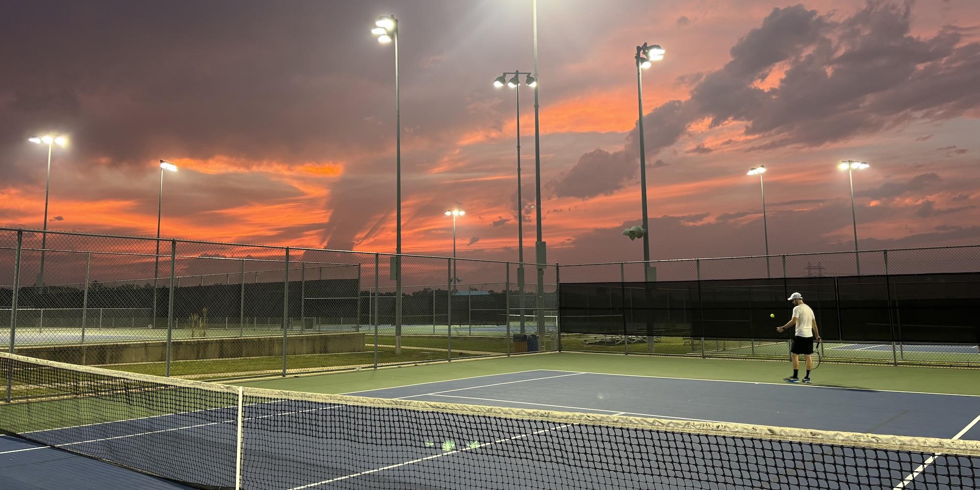 Cover image for this post: A tennis court in the foreground, with my opponent warming up his serve. A beautifully colored sunset is in the background, with the court lights already coming on as it gets dark.