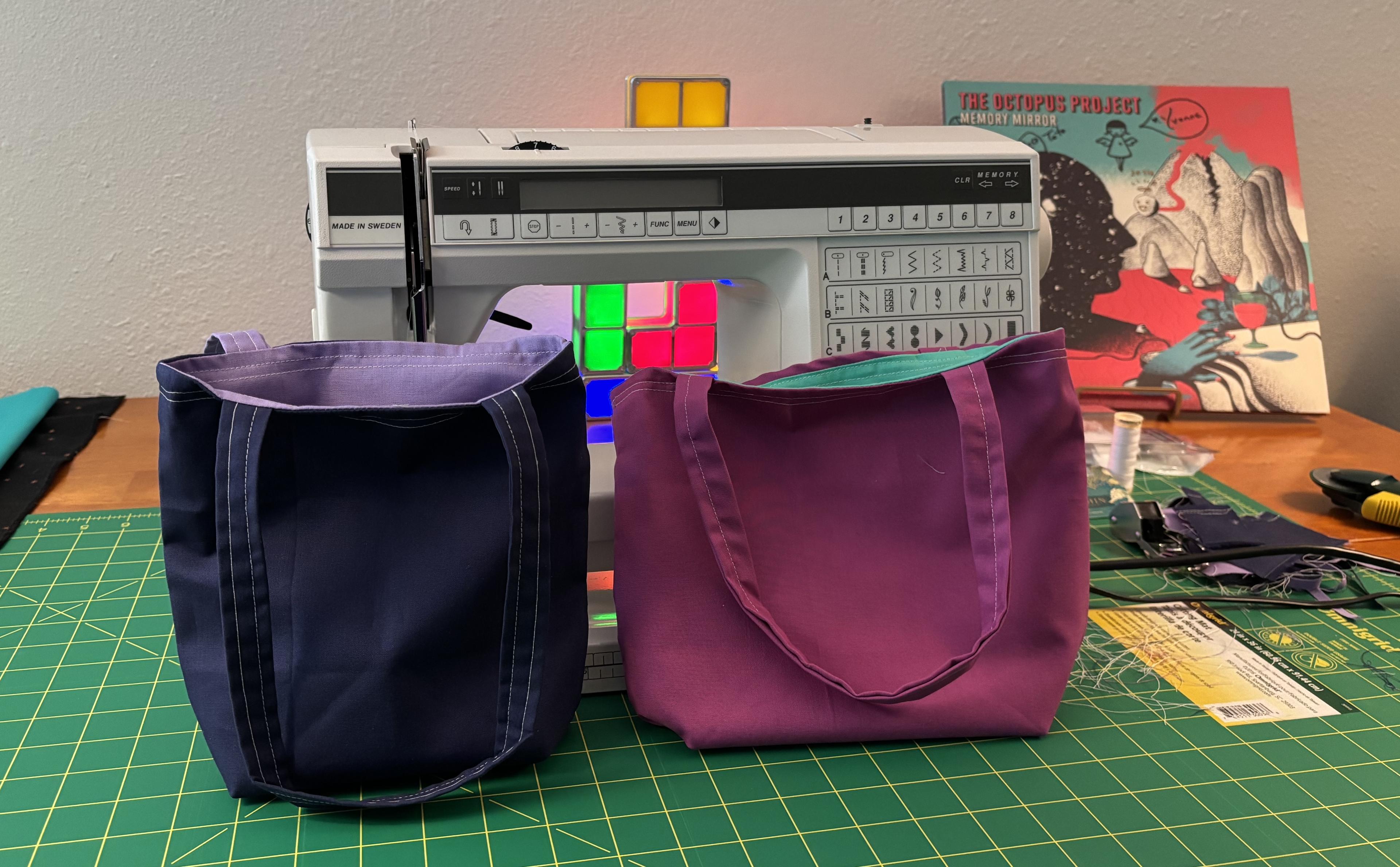Two finished bags sit on my project table in front of the sewing machine. One is dark blue with a lavender lining, and the other is kind of a barney purple with a teal lining. The dark blue bag is visibly better-made than the purple one.