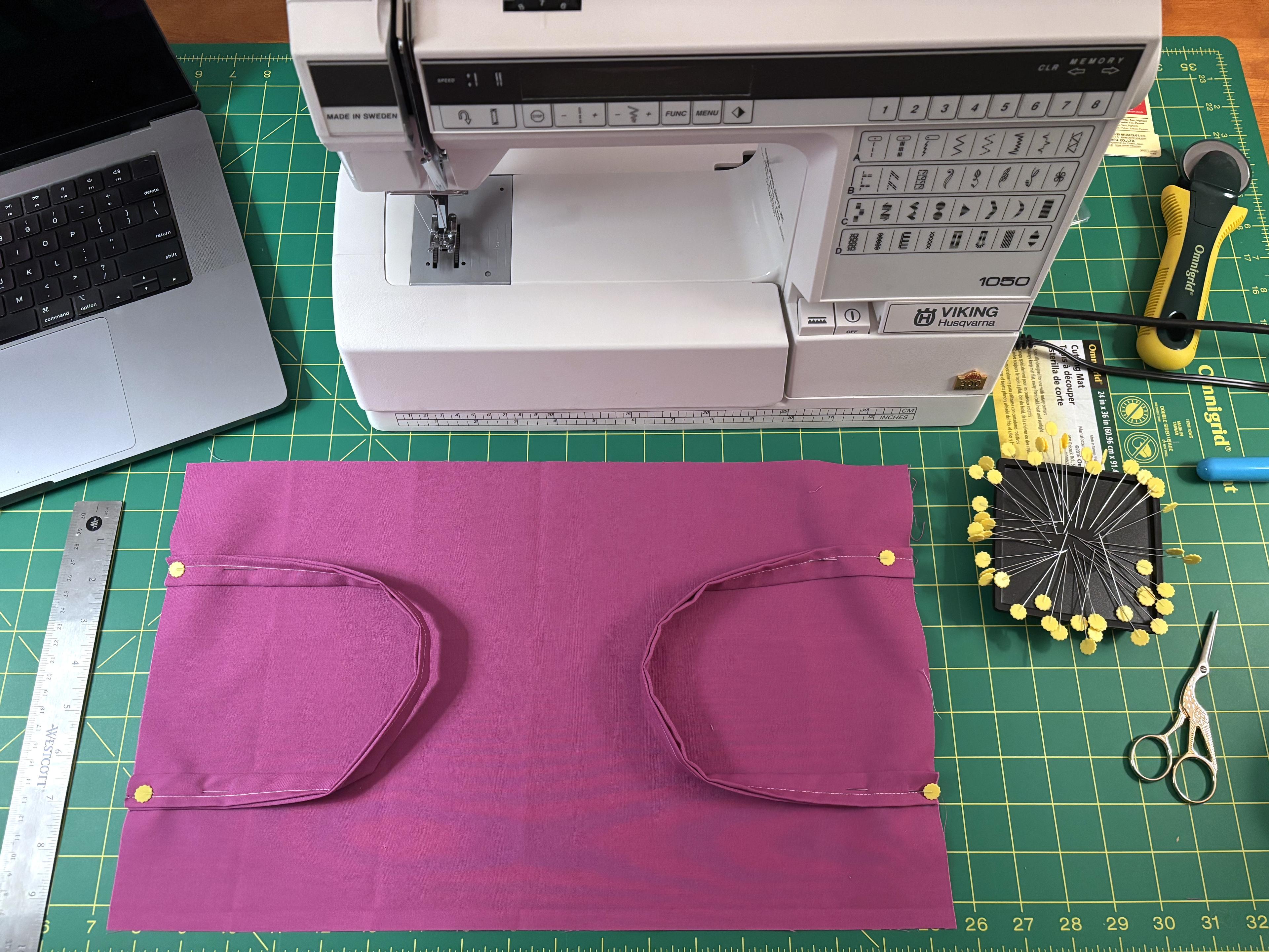 The in-progress tote bag sits on my project table on top of my cutting mat. Right now it's just a rectangle of fabric with the handles pinned in place. Also visible is the corner of my computer, part of my sewing machine, and various bits and bobs like pins, a rotary cutter, and stork scissors.