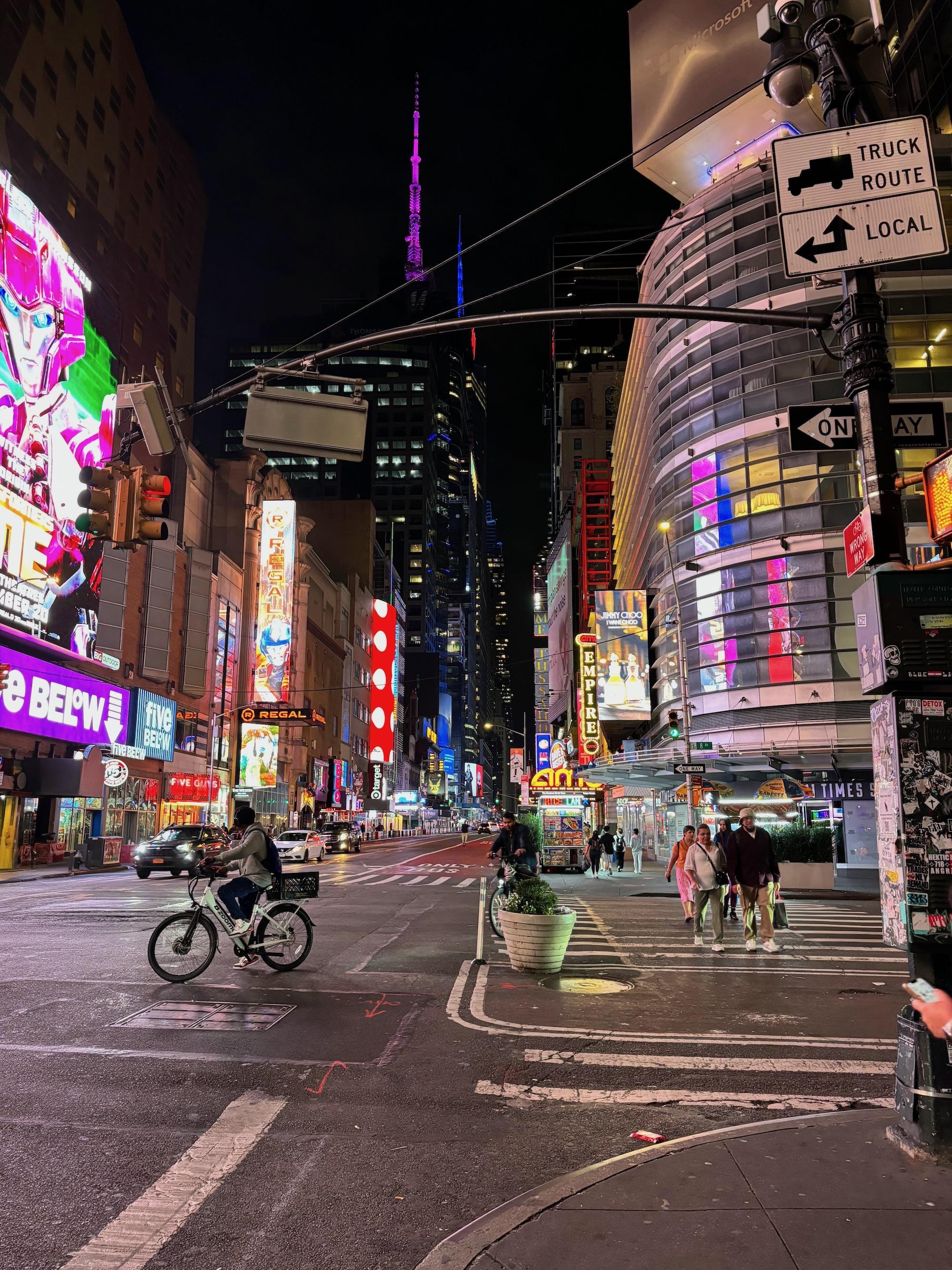 New York City at night. People are biking and walking, and the street is lit by lights and billboards.