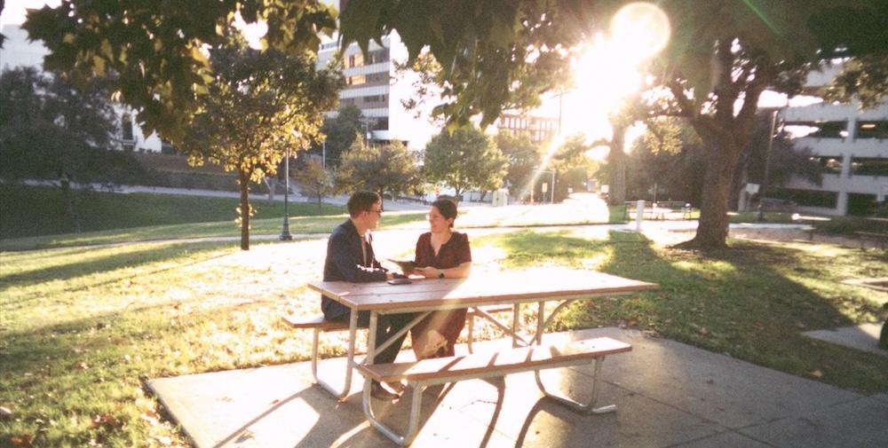 Cover image for this post: Myself and my husband sitting at a picnic table outdoors, with beautiful sunlight.